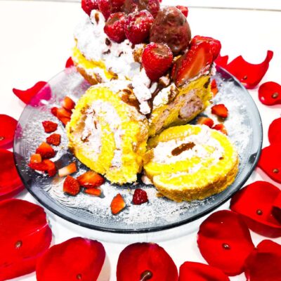 A side view of a gluten free swiss roll that is placed on a blue plate and surrounded by rose petals, while being topped with strawberries, raspberries and chocolate