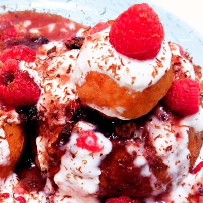 A side top down view of some cheese doughnuts that are topped with jam, sour cream and raspberries, sitting on a plate