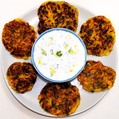 A top down view of 6 zucchini fritters, served on a plate with a bowl of tzatziki sauce