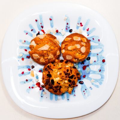 A top down view of some gluten free condensed milk muffins, topped with white chocolate, almond flakes and dried fruits, and served on a plate decorated with a blue liquid